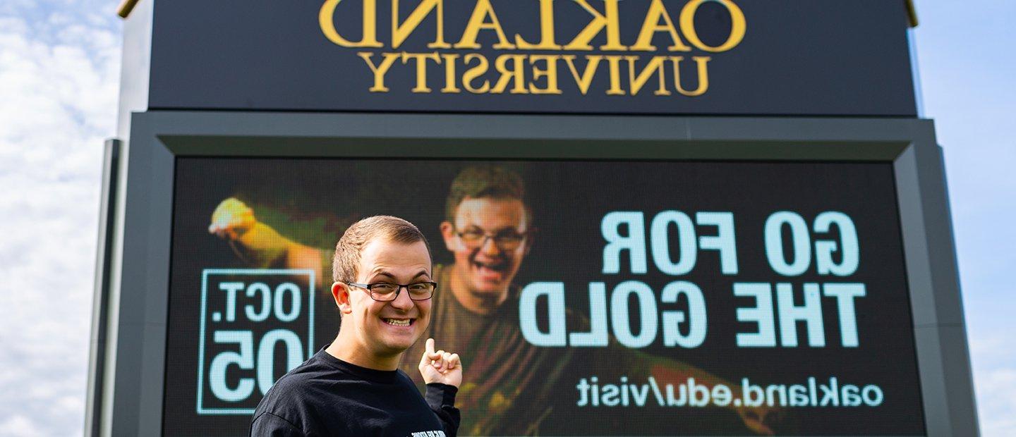 Young man pointing to an ad that he is featured in on an Oakland University electronic marquee.