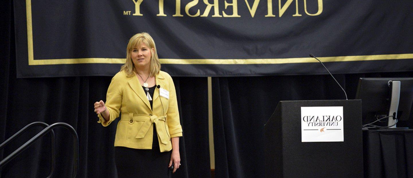 woman speaking on a stage with an Oakland University banner behind her